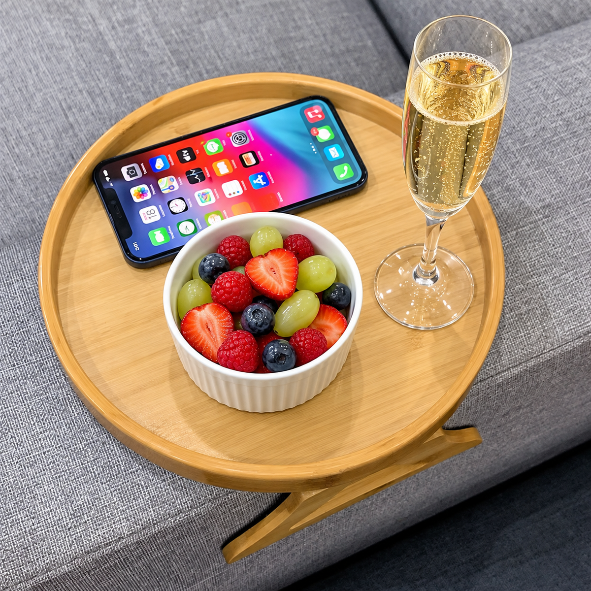 Tablette d'accoudoir NESTIVA en bois posée sur un canapé gris, avec un ramequin de fruits frais et un téléphone